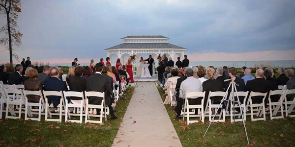 A bride and groom get married in a gazebo overlooking Lake Erie at sunset with wedding guests watching their ceremony